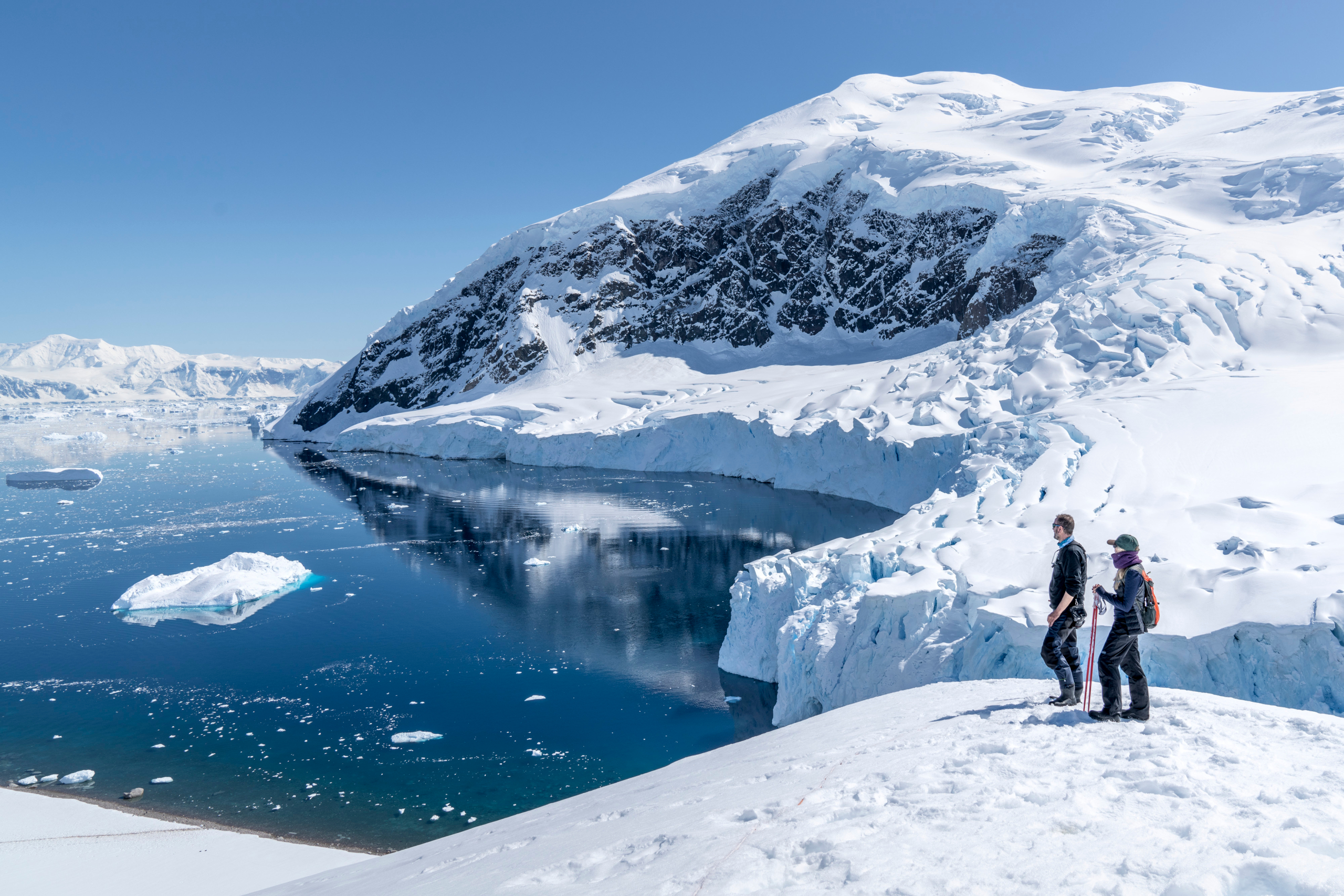 Two guests standing on snowy hillside looking at scenic bay in Antarctica.