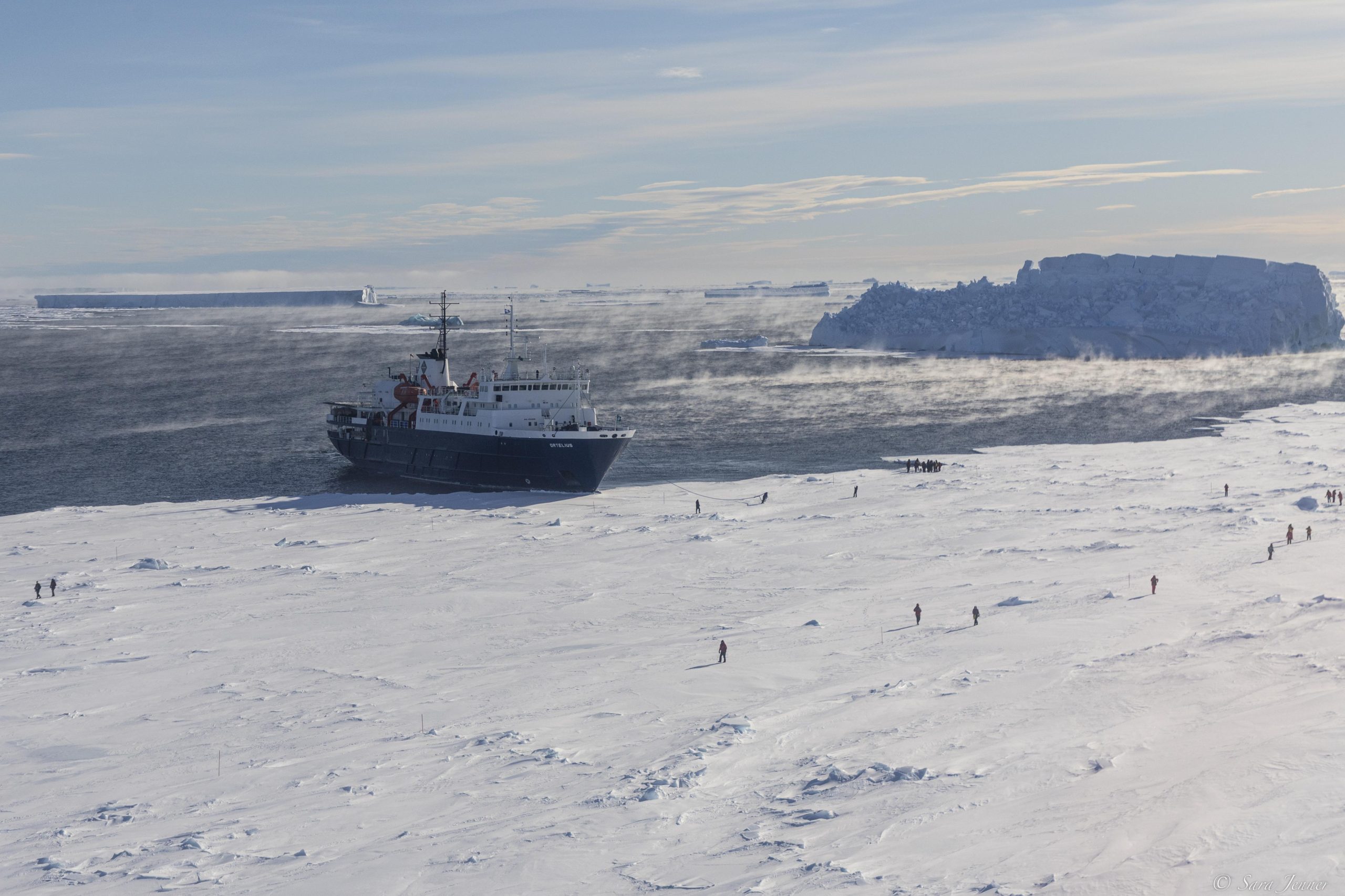 The ship Ortelius anchored at the edge of the fast ice with guests walking on the ice.