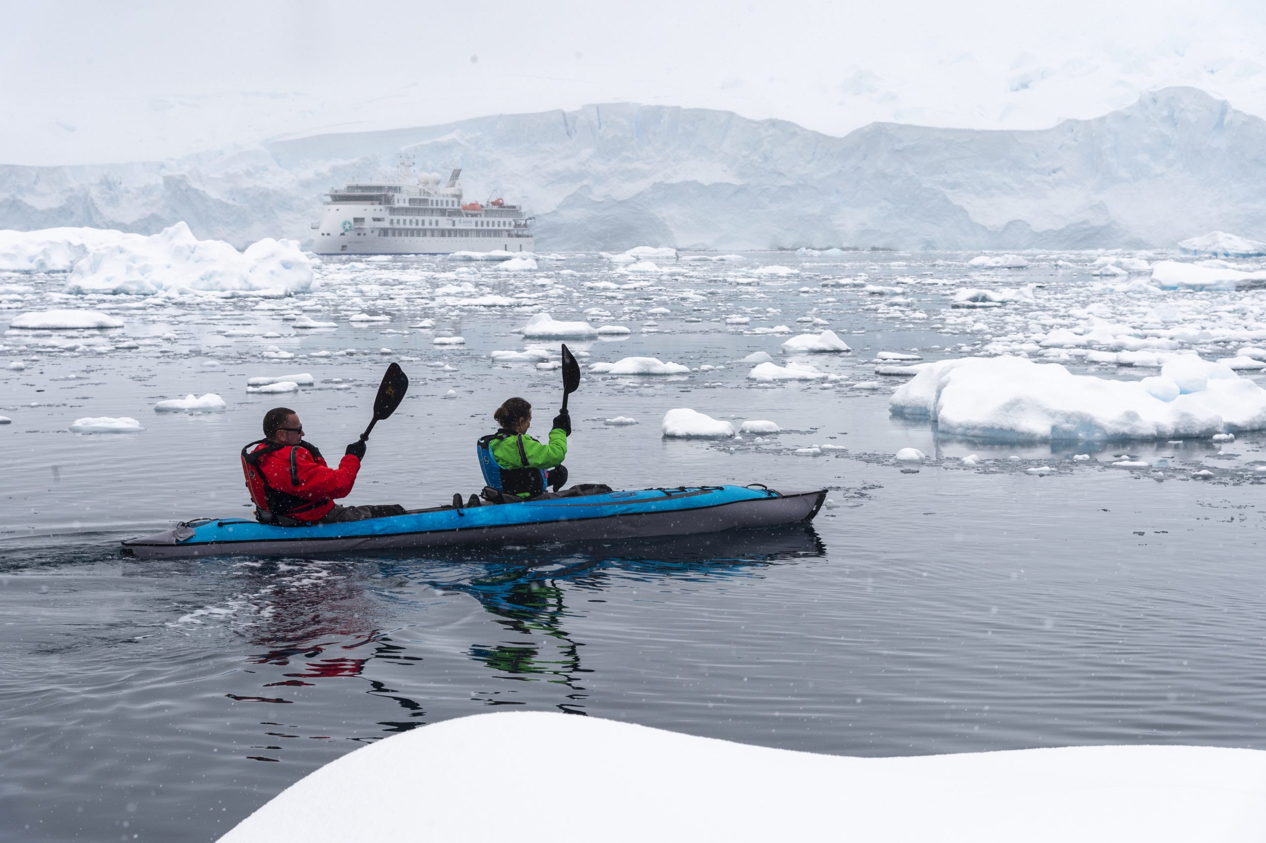 Two guest paddling in icy Antarctic waters with Greg Mortimer ship in the background