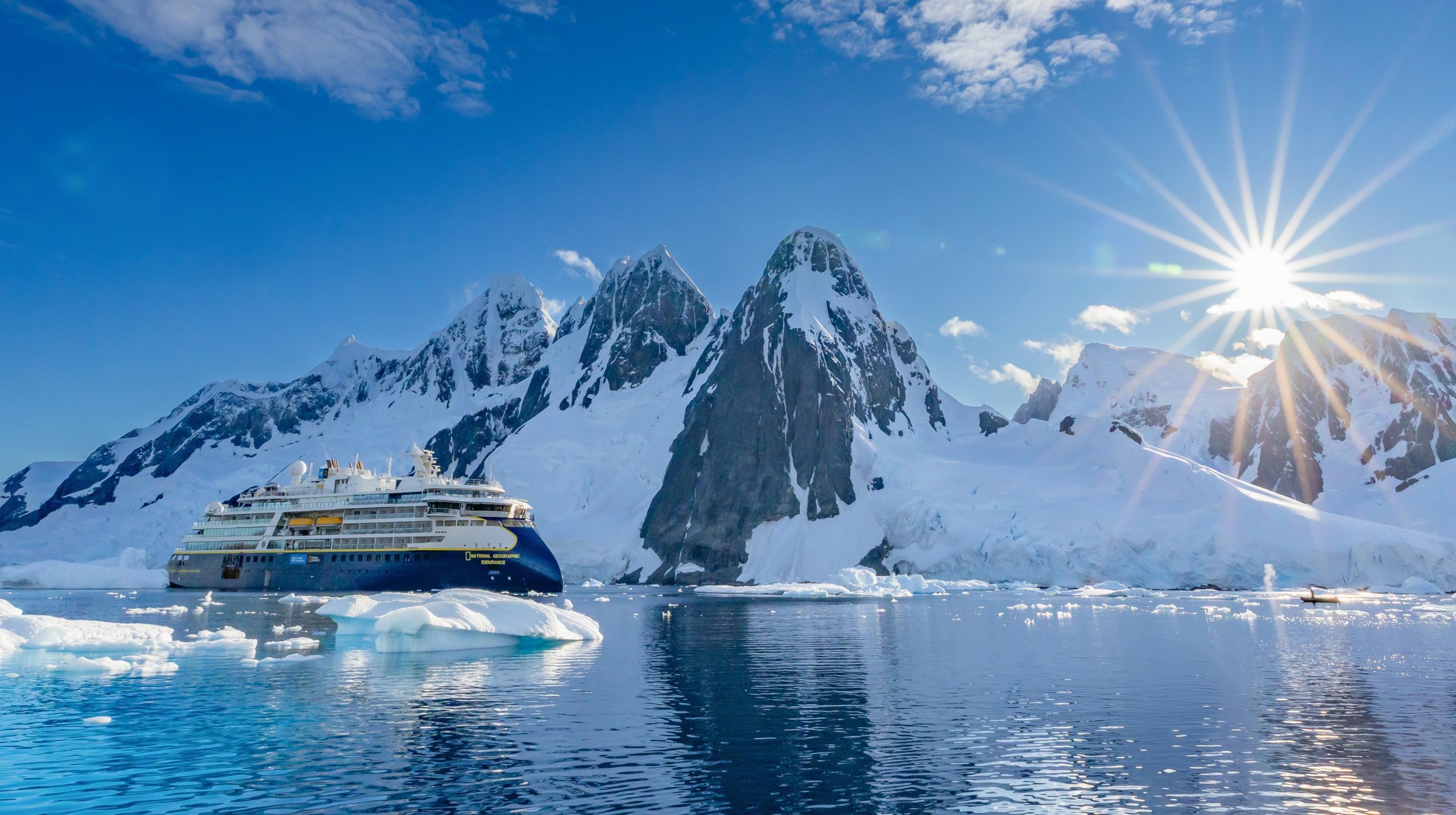 National Geographic ship in narrow channel with towering mountains & sun setting.